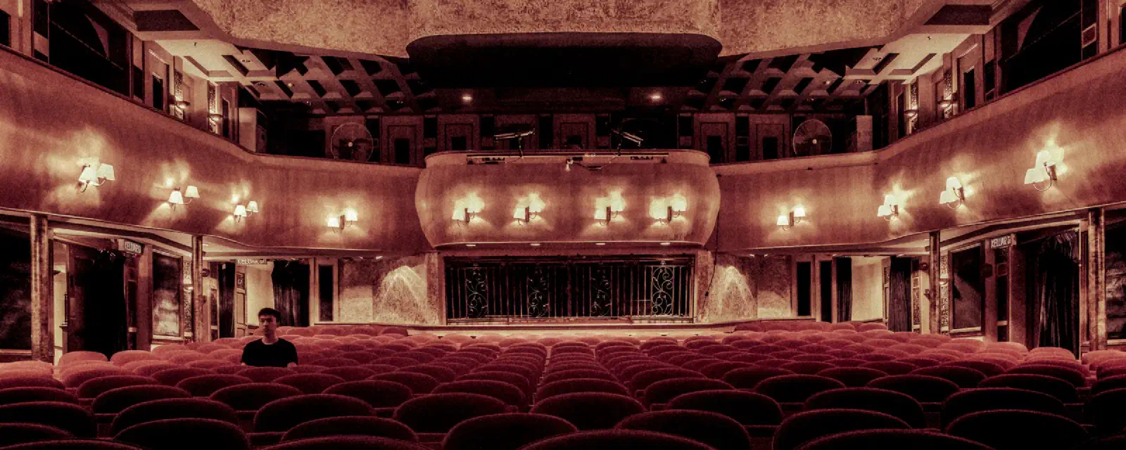En gammel teatersal med rækker af røde fløjlssæder og tændte væglamper.
