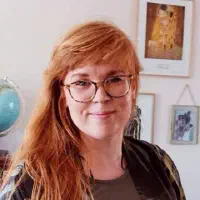 Karen Margrethe S. Kirketerp, smilende, med briller og langt rødt hår, foran en væg med indrammede kunstplakater.